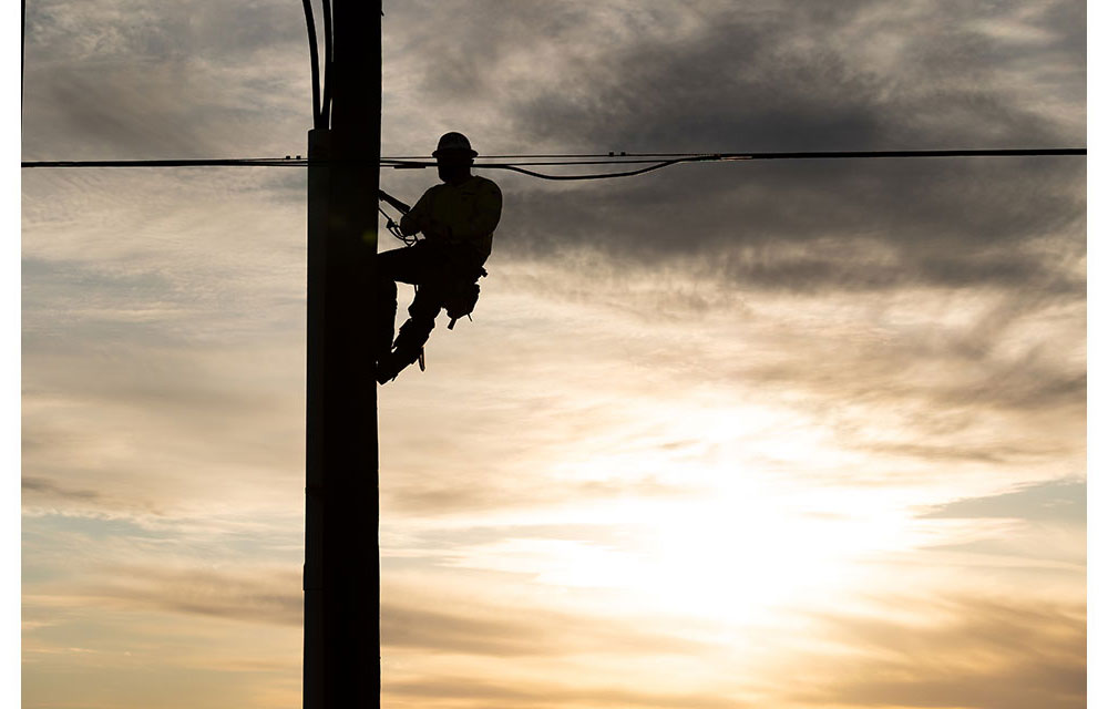 Silhouette of a lineman climbing a utility pole at sunset, secured with safety gear against a cloudy sky.